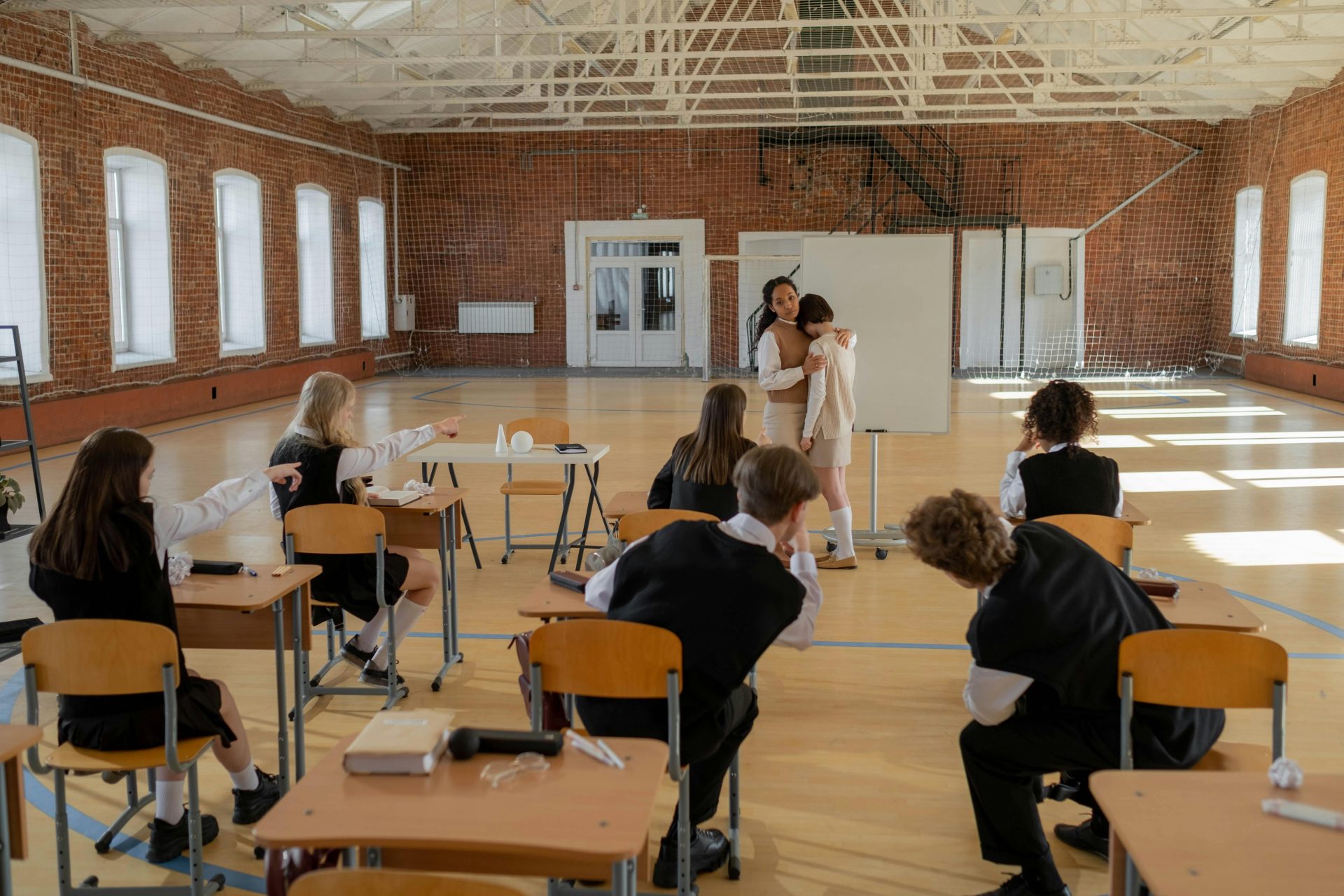 Ein Lehrer tröstet einen Schüler in einem Klassenzimmer, während andere Schüler zusehen. Das Bild fängt die Anspannung und den Zusammenhalt im Schulalltag ein.