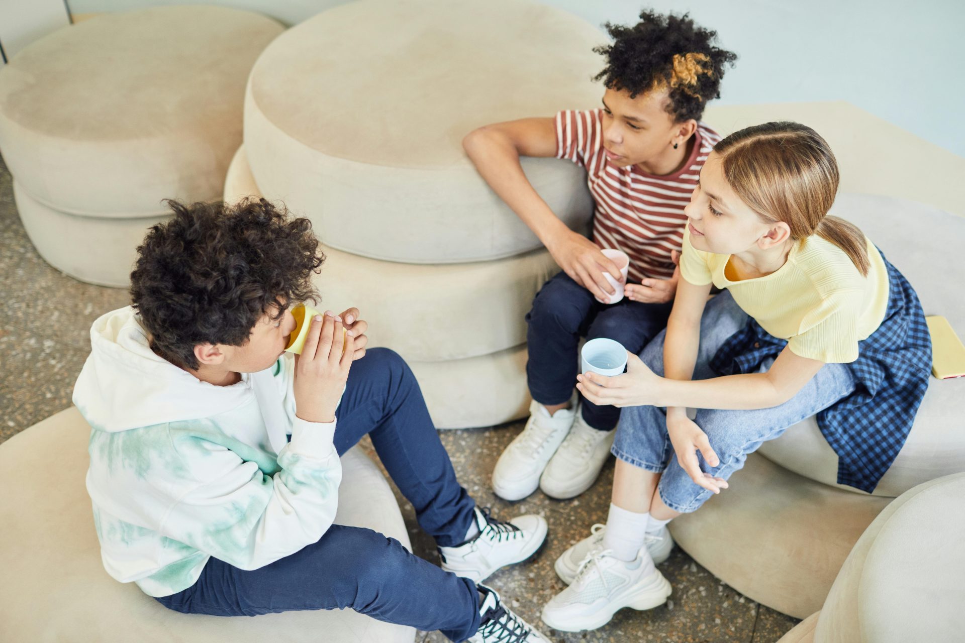 Blick von oben auf eine Gruppe multikultureller Kinder, die auf weichen Kissen sitzen, ein heißes Getränk trinken und gemeinsam Zeit im Wohnzimmer verbringen.