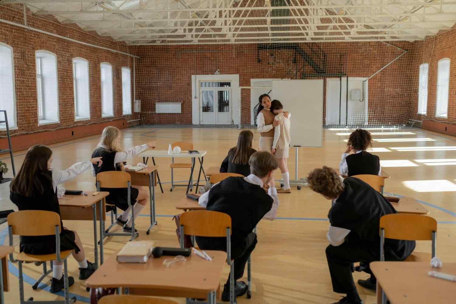 Ein Lehrer tröstet einen Schüler in einem Klassenzimmer, während andere Schüler zusehen. Das Bild fängt die Anspannung und den Zusammenhalt im Schulalltag ein.
